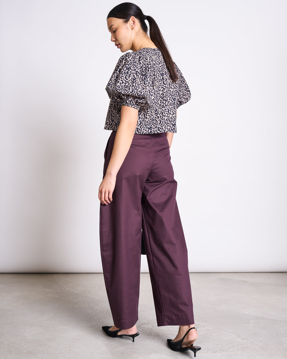 A woman faces away, wearing the jan-n-june CROPPED ORGANIC COTTON BLOUSE OLMOS LEO PRINT with high-waisted plum wide-leg pants and black heels. Her hair is styled in a low ponytail, creating a sustainable look against a light background.