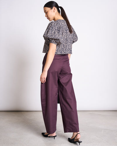 A woman faces away, wearing the jan-n-june CROPPED ORGANIC COTTON BLOUSE OLMOS LEO PRINT with high-waisted plum wide-leg pants and black heels. Her hair is styled in a low ponytail, creating a sustainable look against a light background.