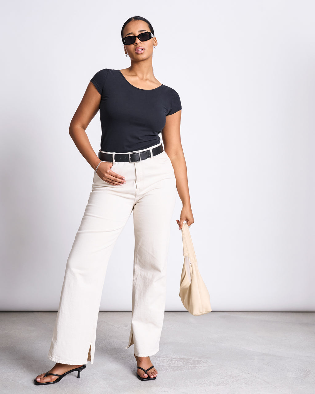 A woman in sunglasses wears the jan-n-june BALLERINA T-SHIRT ALOE SOFT BLACK GOTS with white wide-leg pants, a black belt, black heeled sandals, and holds a beige handbag as she poses confidently against a plain background.