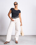 A woman in sunglasses wears the jan-n-june BALLERINA T-SHIRT ALOE SOFT BLACK GOTS with white wide-leg pants, a black belt, black heeled sandals, and holds a beige handbag as she poses confidently against a plain background.