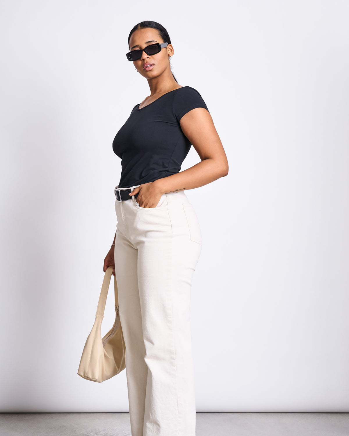 A woman in sunglasses wears the jan-n-june BALLERINA T-SHIRT ALOE SOFT BLACK GOTS with high-waisted cream pants and holds a light beige handbag, standing against a plain white background.