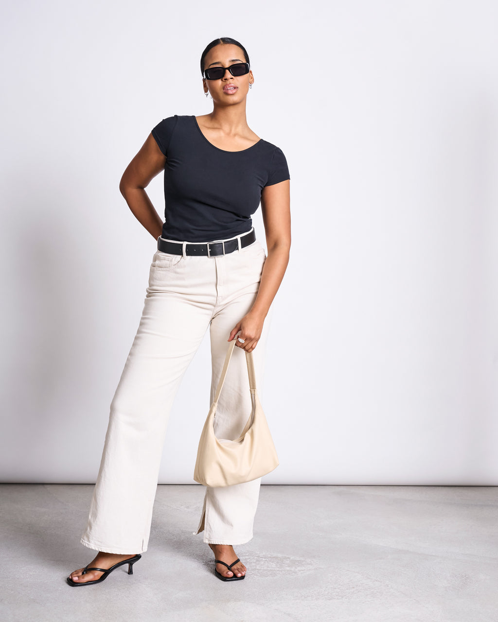 A woman poses confidently in the jan-n-june BALLERINA T-SHIRT ALOE SOFT BLACK GOTS, paired with cream wide-leg pants, a black belt, black heeled sandals, sunglasses, and a beige handbag against a plain backdrop.
