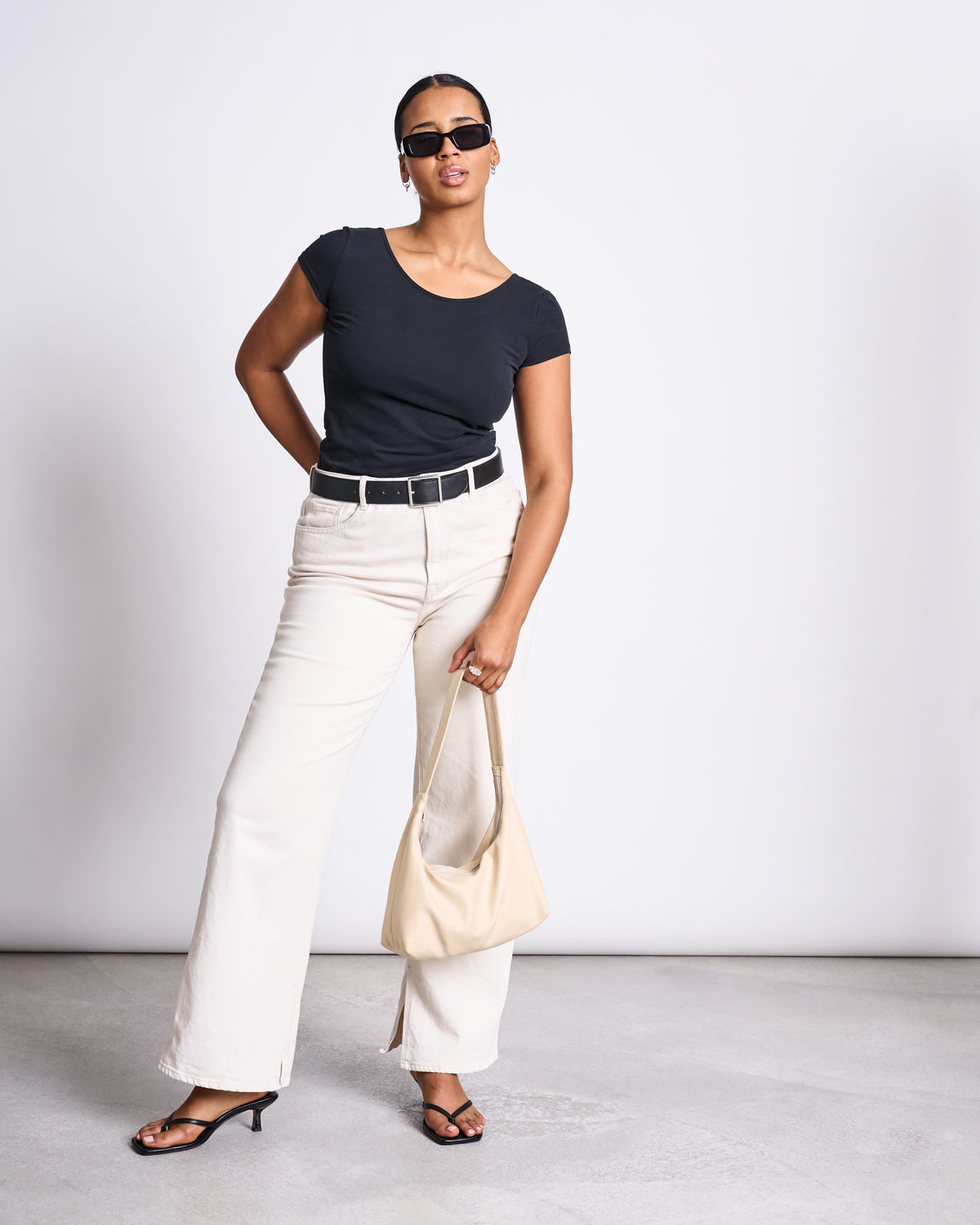 A woman poses confidently in the jan-n-june BALLERINA T-SHIRT ALOE SOFT BLACK GOTS, paired with cream wide-leg pants, a black belt, black heeled sandals, sunglasses, and a beige handbag against a plain backdrop.