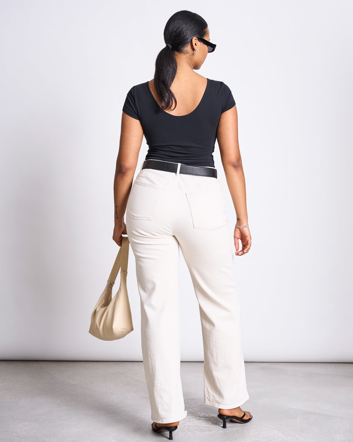 A woman with a low ponytail faces away, wearing the jan-n-june BALLERINA T-SHIRT ALOE SOFT BLACK GOTS, white high-waisted pants, black heeled sandals, sunglasses, and holding a beige handbag.