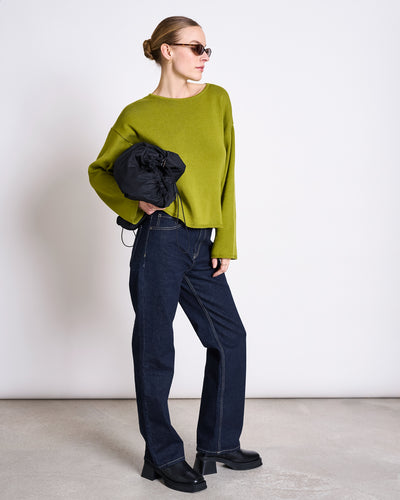 A woman in jan-n-junes LIGHT KNIT JUMPER WILLIAMSBURG MOSSY PEAR GOTS, dark jeans, black boots, and sunglasses stands against a plain background with a black bag—an ideal capsule wardrobe look.