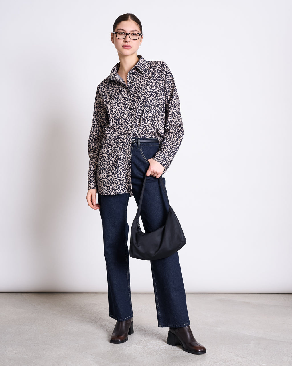 A woman in the jan-n-june OVERSIZED SHIRT MAMRO LEO PRINT, glasses, dark blue jeans, and brown ankle boots stands against a plain white wall, holding a black handbag by her side.