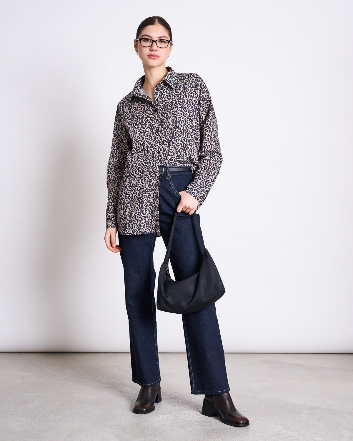 A woman in the jan-n-june OVERSIZED SHIRT MAMRO LEO PRINT, glasses, dark blue jeans, and brown ankle boots stands against a plain white wall, holding a black handbag by her side.