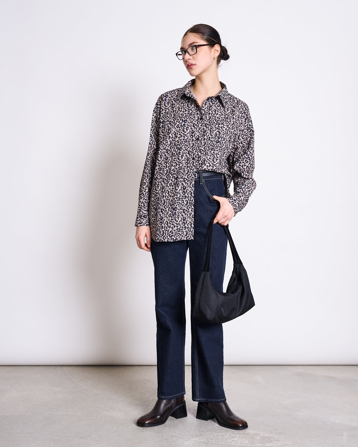 A woman poses against a plain background wearing glasses, the jan-n-june OVERSIZED SHIRT MAMRO LEO PRINT, dark high-waisted jeans, black ankle boots, and carrying a black shoulder bag.