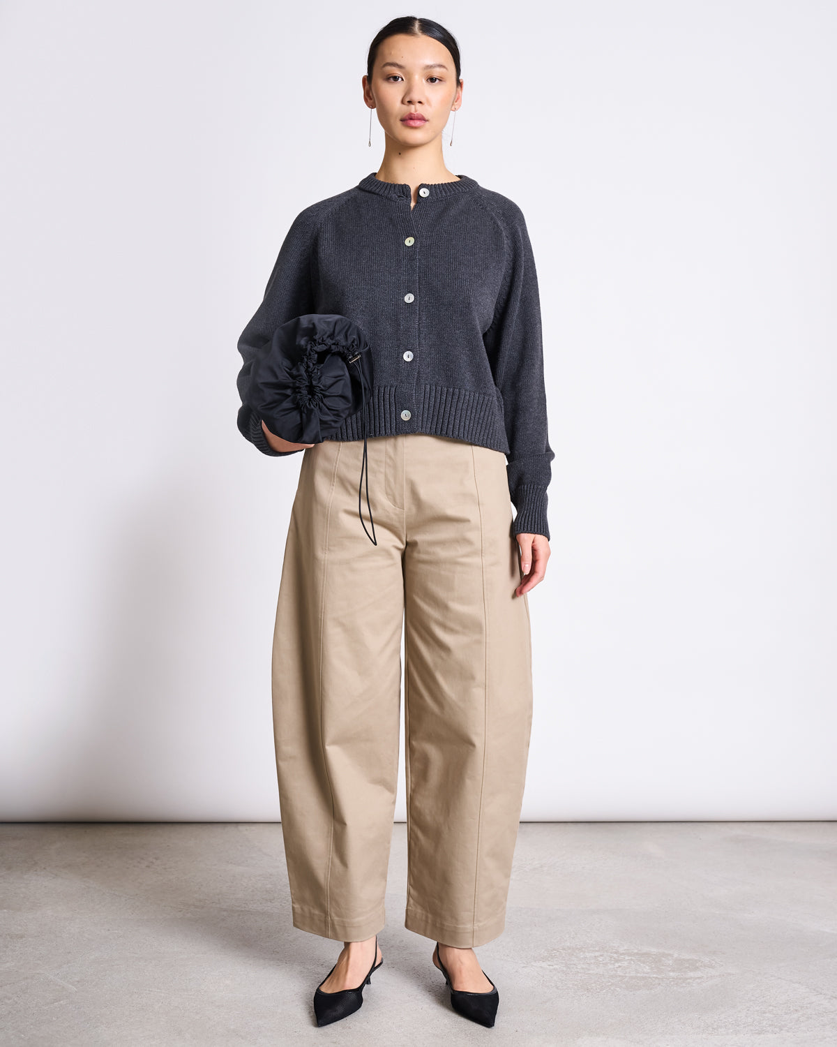 A woman stands against a plain background wearing the jan-n-june RELAXED CREWNECK CARDIGAN SIVIA ANTHRACITE GOTS, paired with beige wide-leg pants and black pointed shoes, holding a dark gathered item in her left hand.