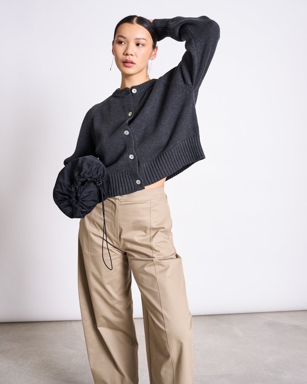 A woman poses against a plain background, wearing the jan-n-june RELAXED CREWNECK CARDIGAN SIVIA ANTHRACITE GOTS and beige wide-leg pants, holding a black ruched handbag with her left hand resting on her head.