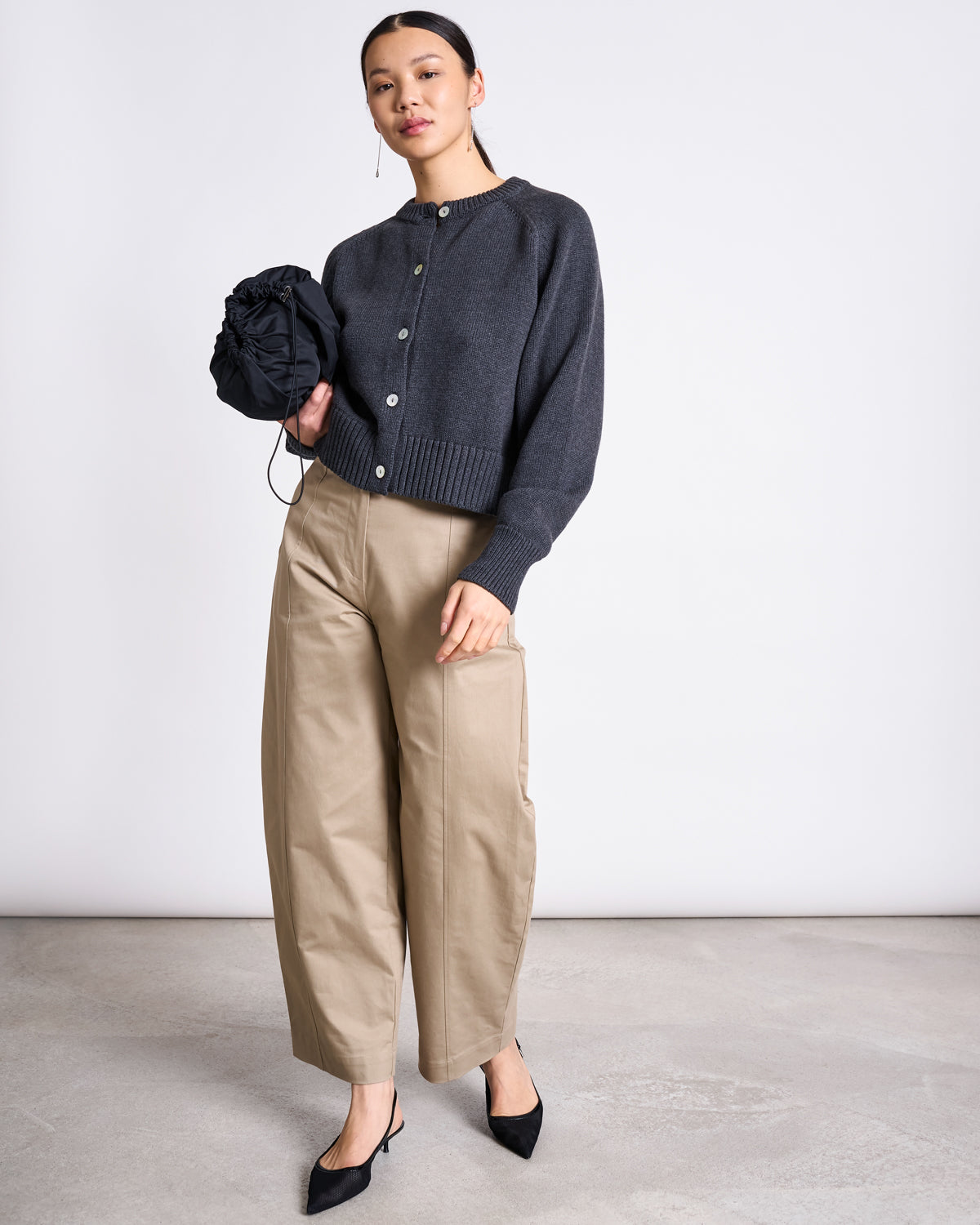 A woman stands against a plain background in the jan-n-june RELAXED CREWNECK CARDIGAN SIVIA ANTHRACITE GOTS, beige wide-leg pants, black pointed shoes, and holds a black scrunched bag with her hair pulled back.