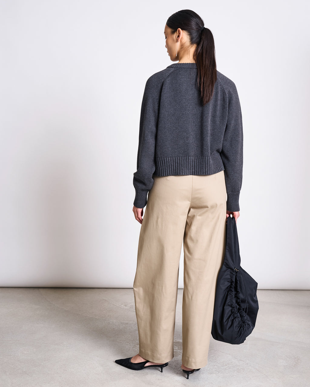 A woman with long dark hair in a ponytail stands facing away, wearing the jan-n-june RELAXED CREWNECK CARDIGAN SIVIA ANTHRACITE GOTS, beige wide-leg pants, black pointed shoes, and holding a black bag in her left hand.