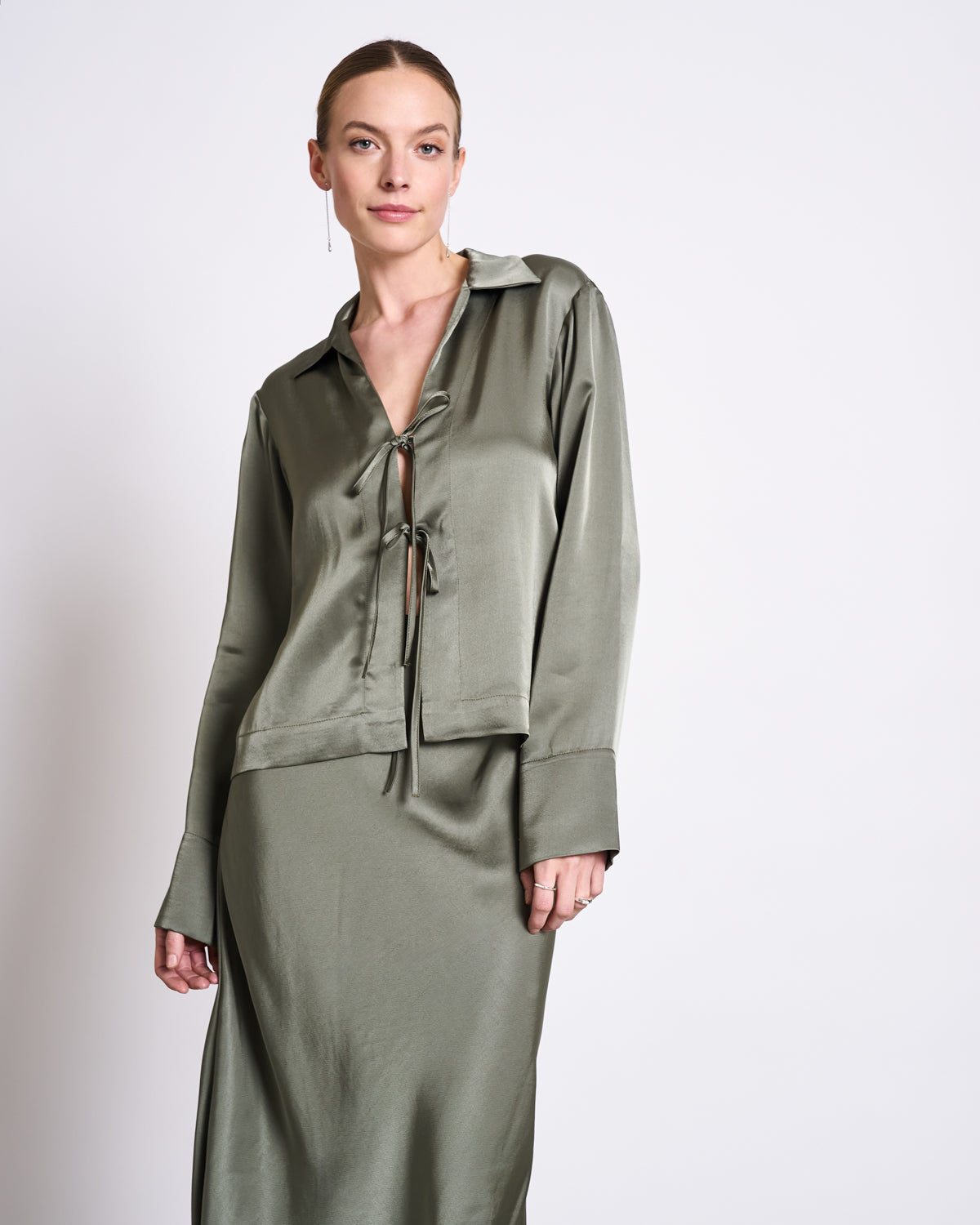 A woman with her hair pulled back and a neutral expression wears the jan-n-june TIE BLOUSE KALUZ SEA SPRAY, a silky sage green vegan silk blouse with a tie front, paired with a matching satin skirt against a plain white background.