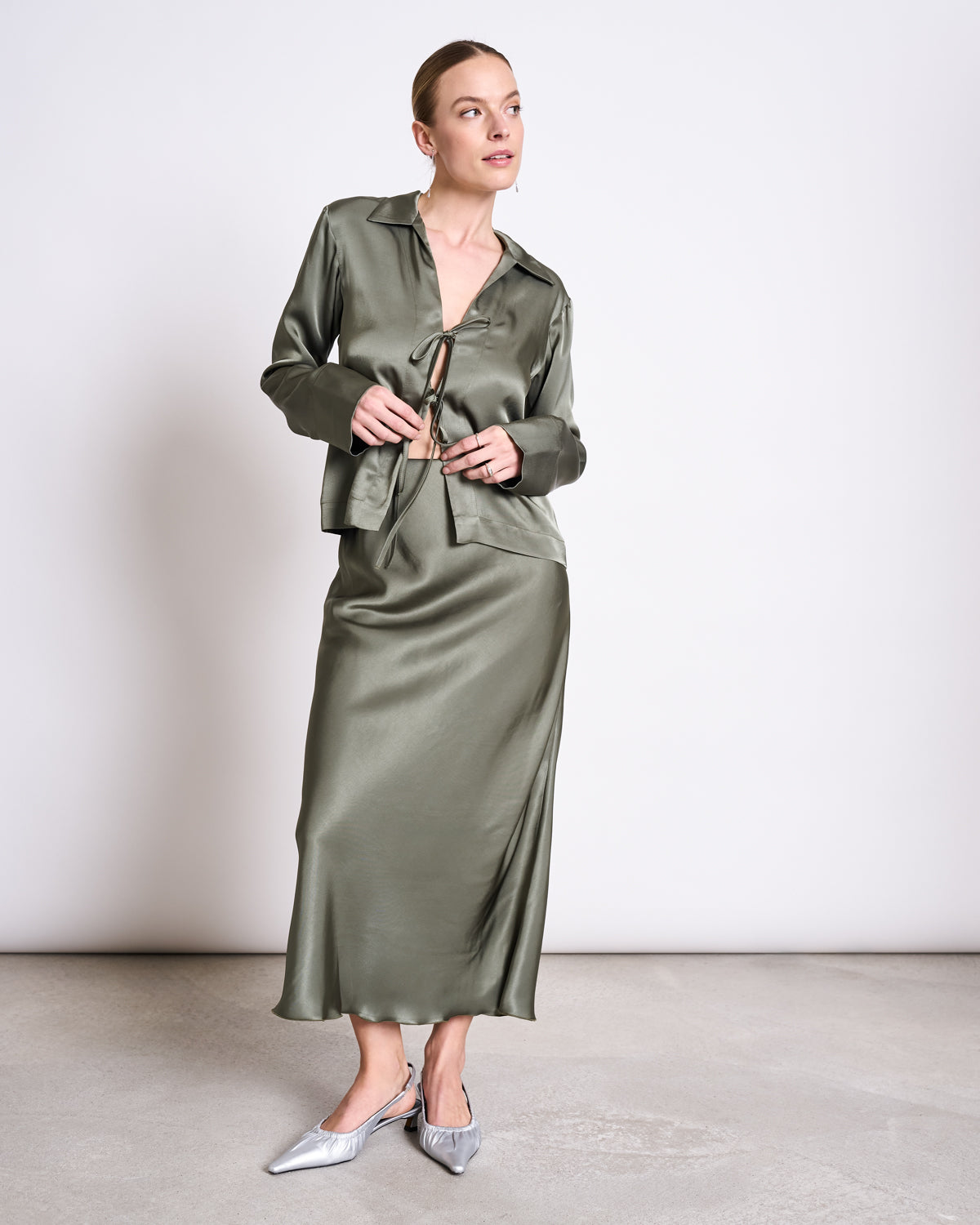 A woman stands indoors wearing the jan-n-june TIE BLOUSE KALUZ SEA SPRAY, a shiny sage green vegan silk top with a tie front and matching skirt. She pairs it with silver pointed-toe heels, her hair pulled back as she looks to the side.