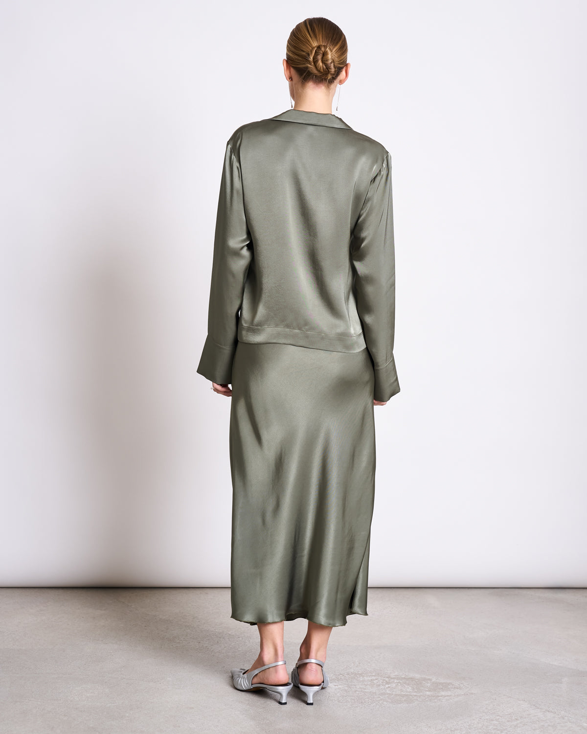 A woman stands facing away in the jan-n-june TIE BLOUSE KALUZ SEA SPRAY, an olive green long-sleeved blouse with a matching midi skirt. Her hair is in a neat bun and she wears silver pointed-toe kitten heels on a light gray floor.