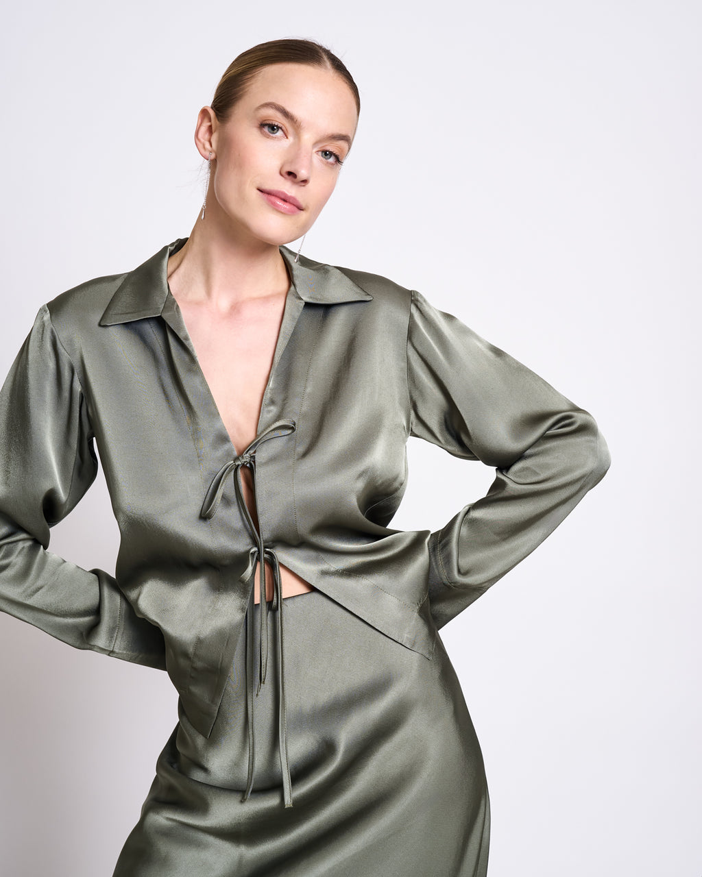 A woman models the jan-n-june TIE BLOUSE KALUZ SEA SPRAY in shiny sage green vegan silk, featuring a deep V-neck and tie detail, paired with a matching skirt, standing against a plain light background.