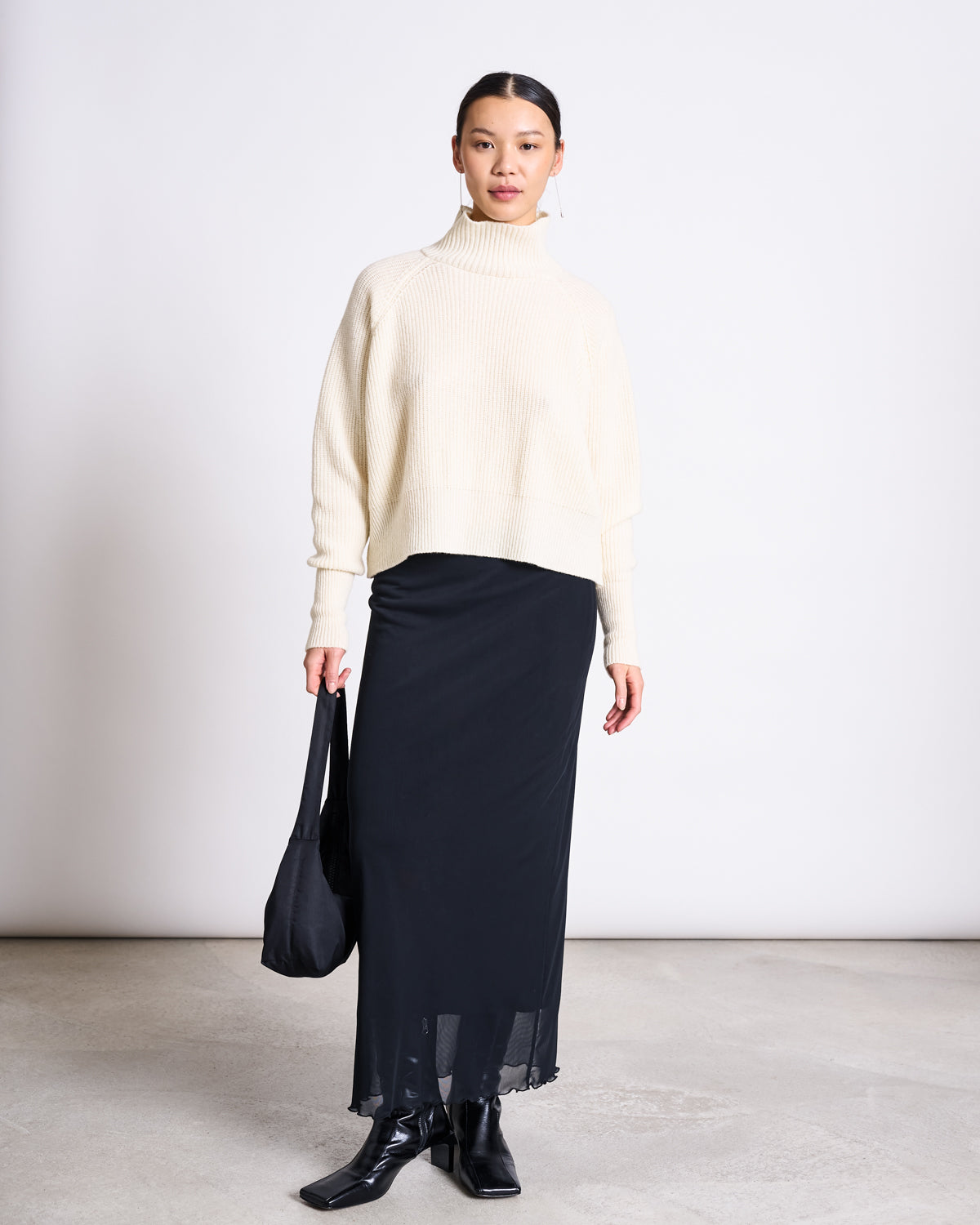 A woman stands against a plain background wearing the jan-n-june HIGH-NECK JUMPER WOOLY OLA OFFWHITE GOTS, paired with a long black skirt, black boots, and holding a black bag in her left hand. She looks toward the camera with a neutral expression.
