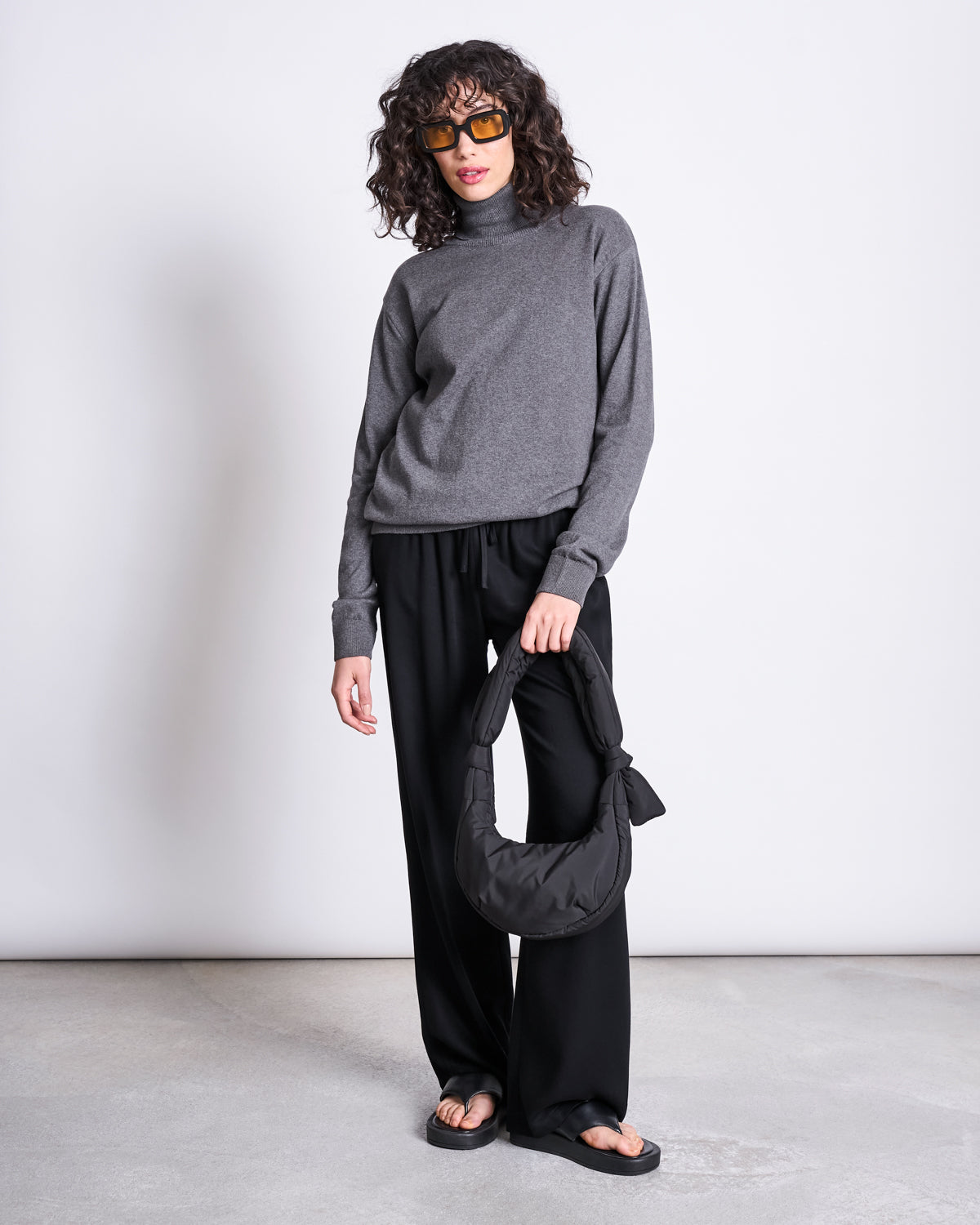 A person with curly hair in sunglasses, a gray turtleneck, loose black pants, and black sandals stands against a plain background holding the jan-n-june BAG BIWA PUFFY MICRO BLACK, an ultralight bag made from recycled polyester.