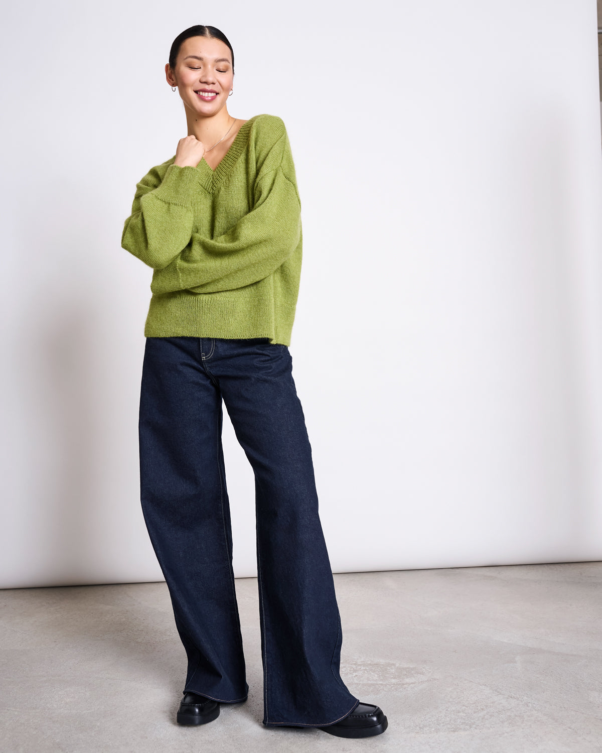 A person smiles in the jan-n-june REC. MOHAIR V-NECK JUMPER YICA MOSSY PEAR, styled with wide-leg dark blue jeans and black shoes, posing against a plain white background.