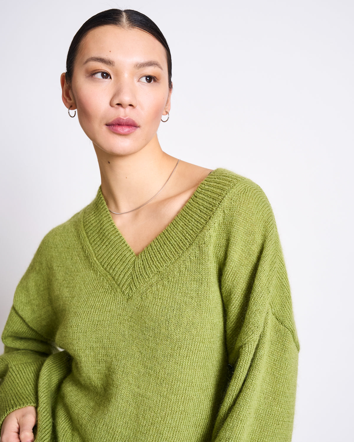 A woman with dark hair pulled back models the jan-n-june REC. MOHAIR V-NECK JUMPER YICA in Mossy Pear, paired with small hoop earrings, standing against a plain white background and embodying a cozy winter look.