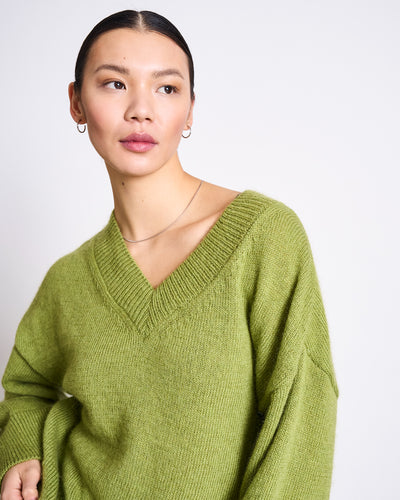 A woman with dark hair pulled back models the jan-n-june REC. MOHAIR V-NECK JUMPER YICA in Mossy Pear, paired with small hoop earrings, standing against a plain white background and embodying a cozy winter look.