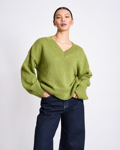A person stands confidently with one hand in their pocket, wearing the jan-n-june REC. MOHAIR V-NECK JUMPER YICA MOSSY PEAR and dark wide-leg jeans against a plain white background.