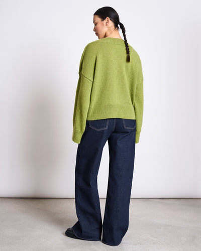 A person with a braided ponytail faces away, wearing the jan-n-june REC. MOHAIR V-NECK JUMPER YICA MOSSY PEAR and wide-leg dark blue jeans against a plain white background.
