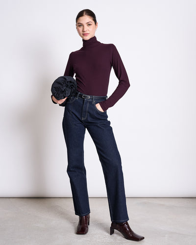 A woman stands against a plain background, smiling slightly at the camera while wearing the jan-n-june LYOCELL TURTLENECK MIO CHERRY LACQUER, paired with dark jeans, a black belt, brown ankle boots, and holding a folded dark jacket or bag.