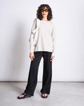 A woman with curly hair and sunglasses wears the jan-n-june BRUSHED LONGSLEEVE BOY IVORY GOTS top, black wide-leg pants, black sandals, and carries a white bag over her shoulder while standing against a plain white background.