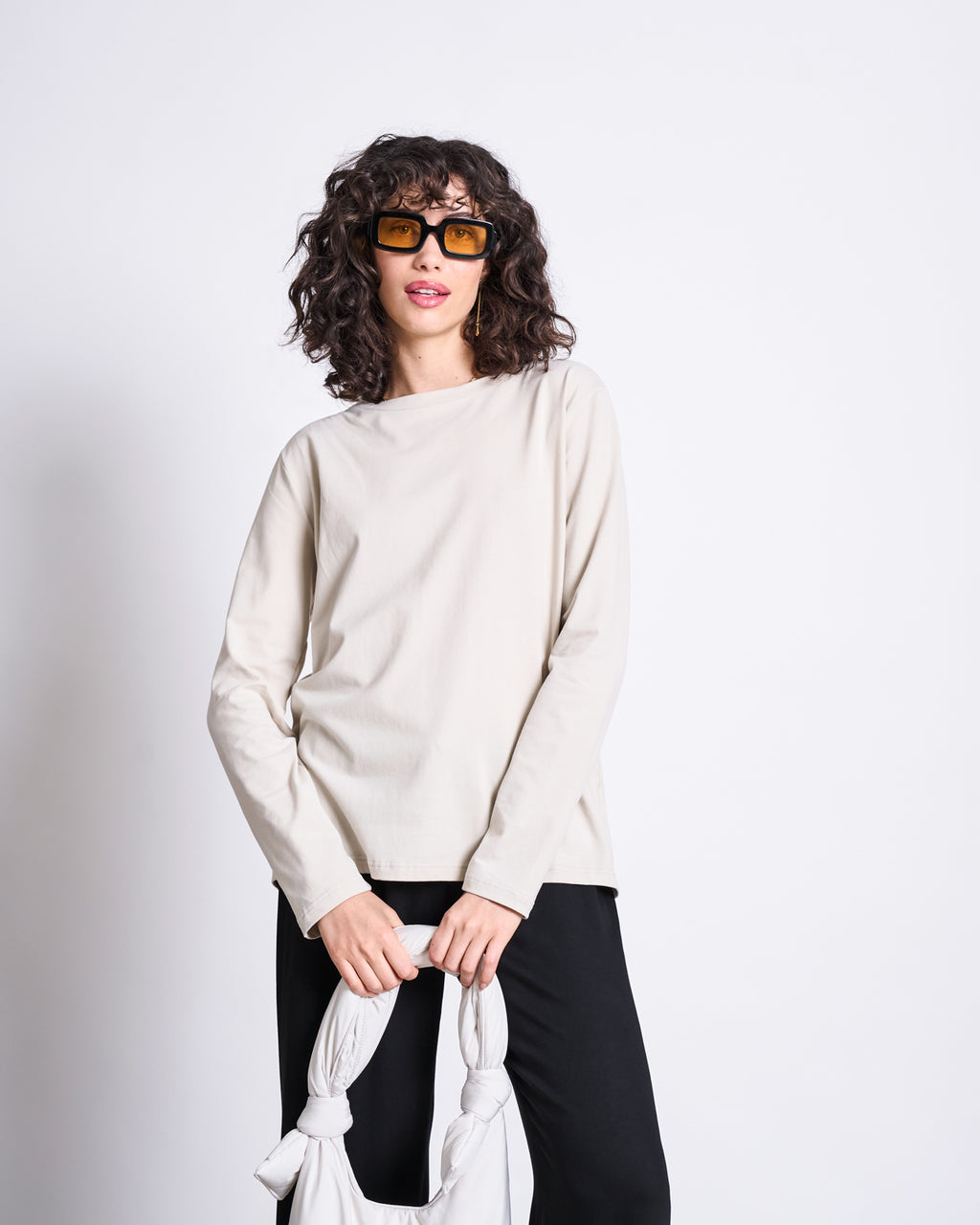 A person with curly brown hair, sunglasses, and black pants stands against a plain background wearing the jan-n-june BRUSHED LONGSLEEVE BOY IVORY GOTS top and holding a white knotted bag.