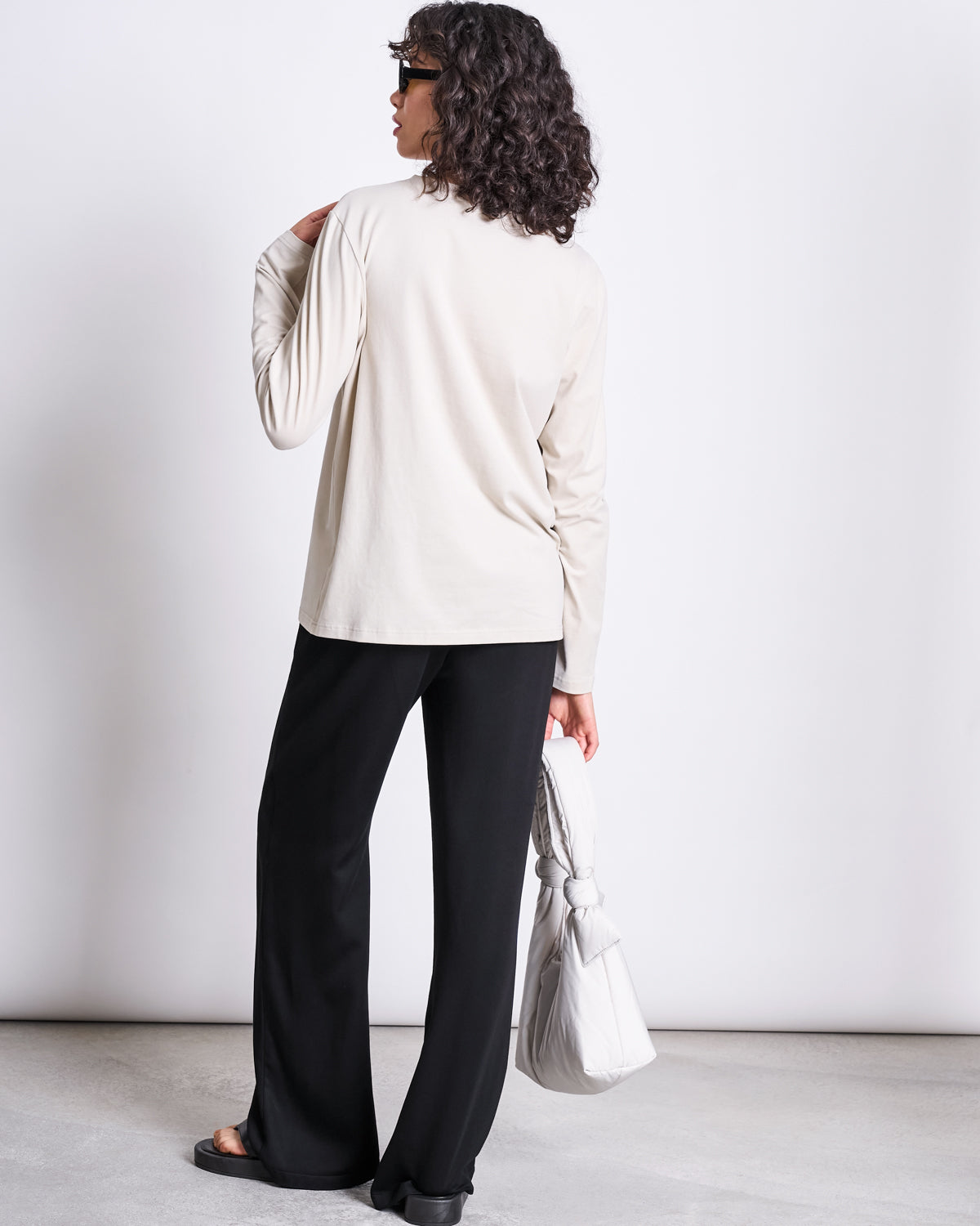 A person with curly hair and sunglasses models the jan-n-june BRUSHED LONGSLEEVE BOY IVORY GOTS top, paired with black wide-leg pants and a large white bag, standing against a plain white background.