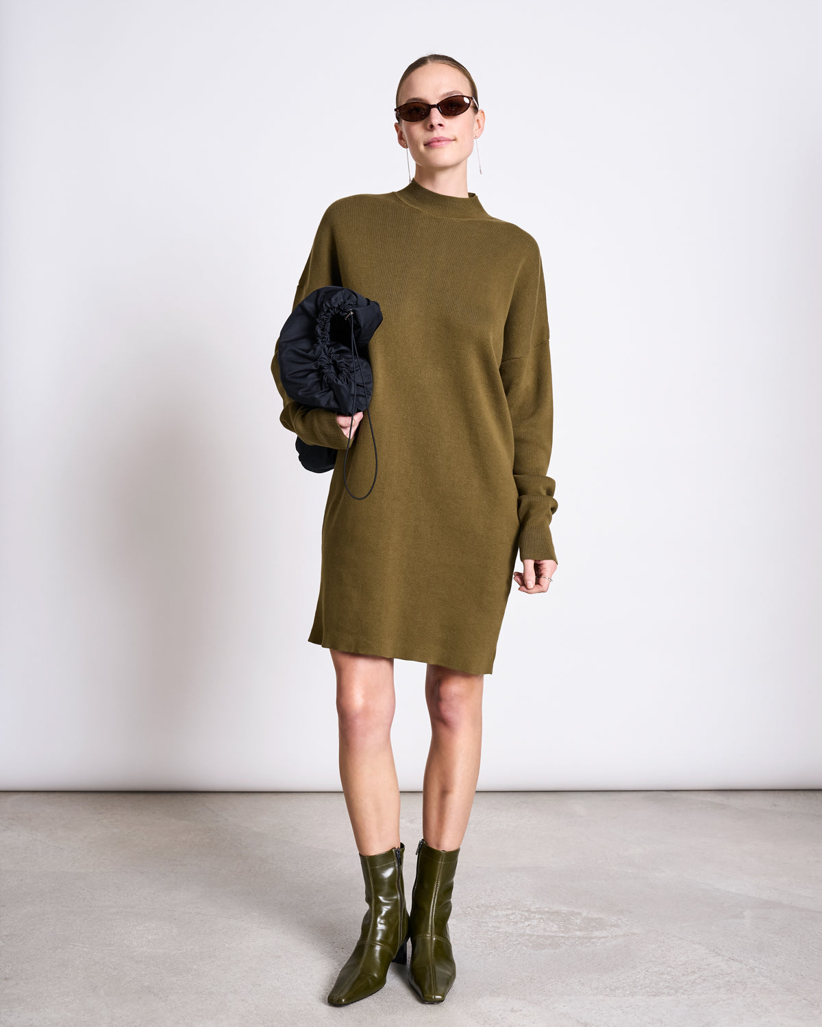 A woman poses against a plain backdrop wearing the KNIT DRESS NUNA WOODY OLIVE GOTS by jan-n-june, paired with matching shiny ankle boots, sunglasses, and holding a black clutch.