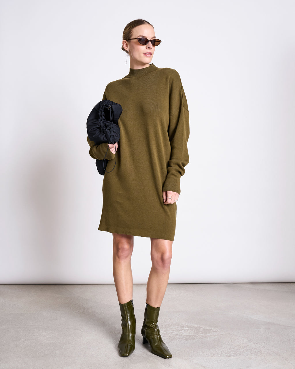 A woman in the jan-n-june KNIT DRESS NUNA WOODY OLIVE GOTS pairs it with matching ankle boots, sunglasses, hoop earrings, and holds a black jacket while standing against a plain white wall.