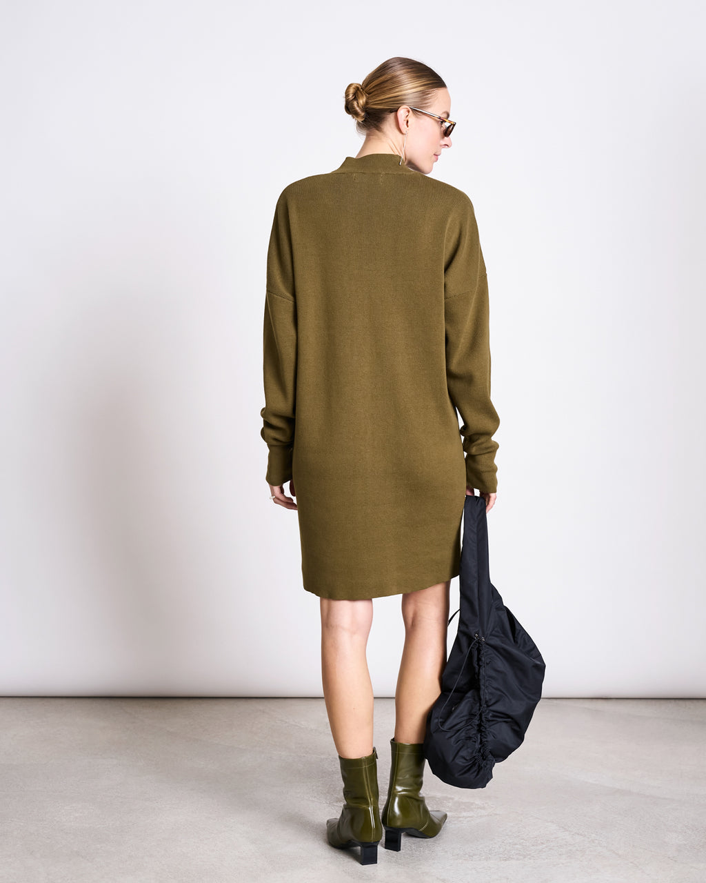 A woman faces away, wearing the jan-n-june KNIT DRESS NUNA WOODY OLIVE GOTS with matching ankle boots and sunglasses. She carries a black bag and has her hair in a bun against a plain, light background.