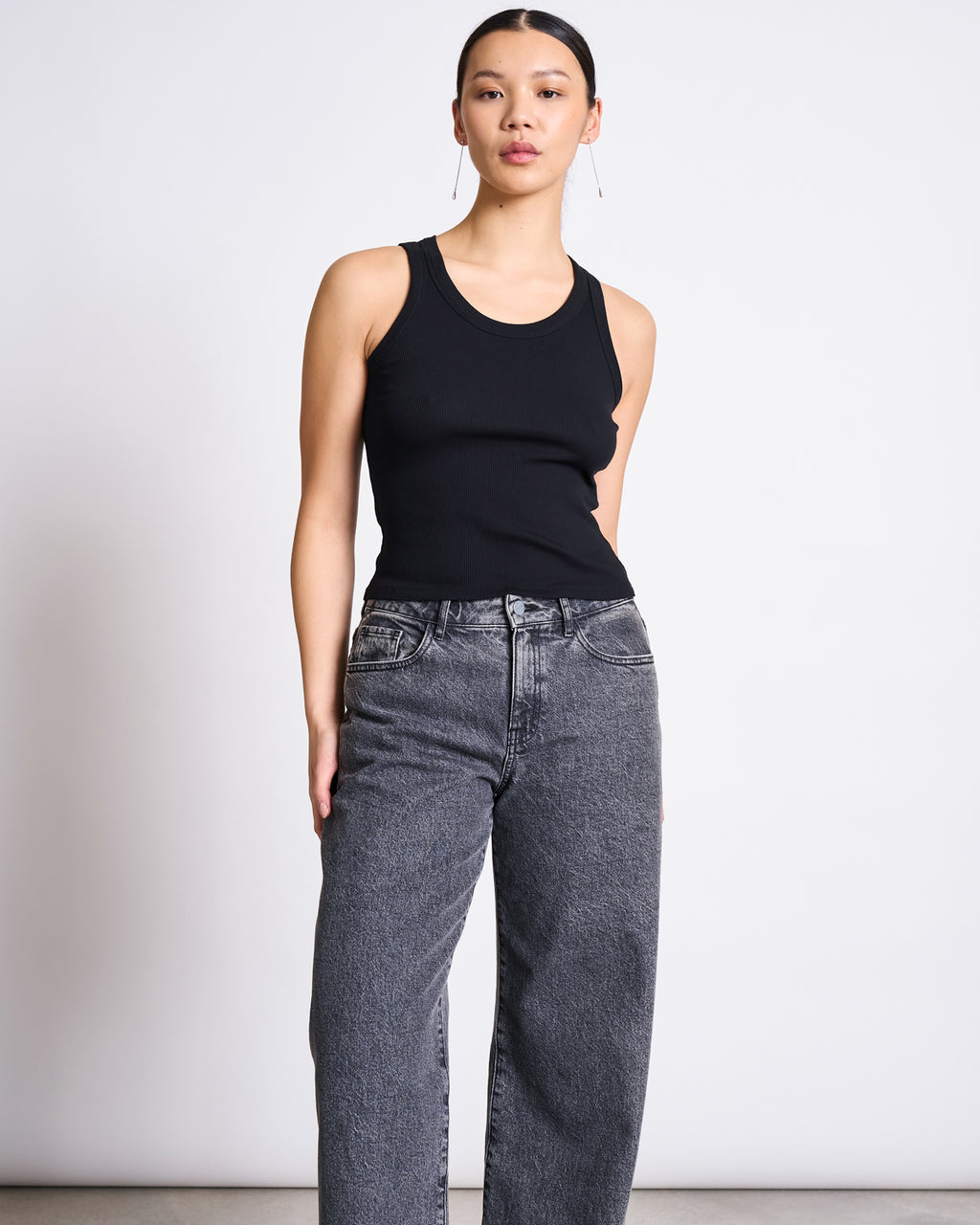 A woman stands against a plain white background, wearing the jan-n-june MINIRIB TANK TOP AMAGER BLACK GOTS and high-waisted, loose-fitting gray jeans, with her hair pulled back and a neutral expression.