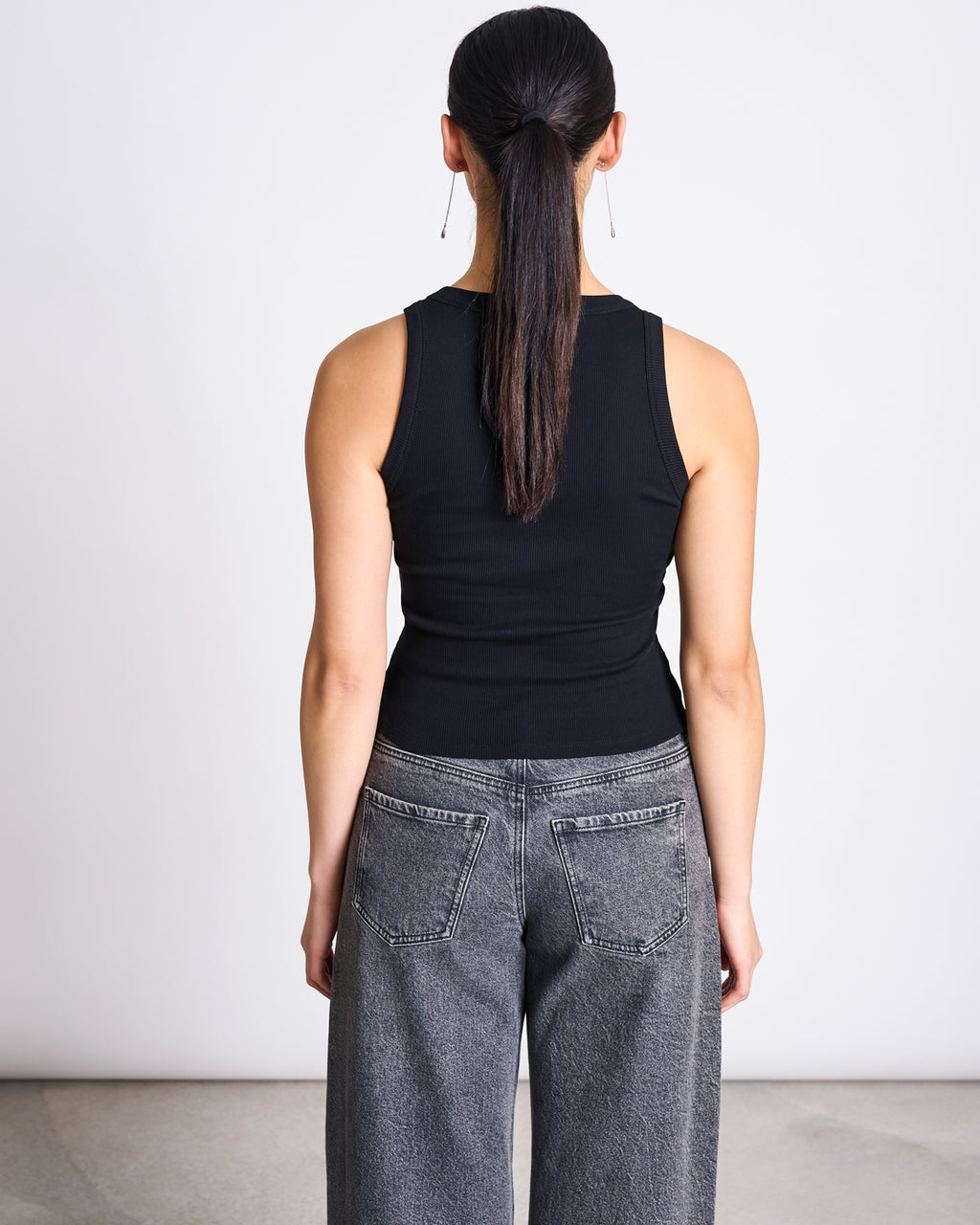A woman with long dark hair in a ponytail stands facing away from the camera, wearing the jan-n-june MINIRIB TANK TOP AMAGER BLACK GOTS and high-waisted gray jeans with large back pockets, against a plain white wall.