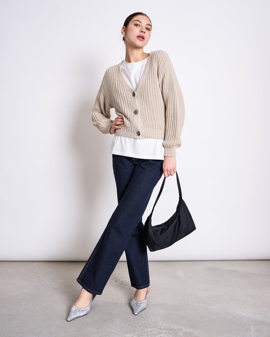 A woman stands against a plain white wall, wearing the jan-n-june CHUNKY CARDIGAN LENA IVORY GOTS over a white top, paired with dark blue jeans, silver pointed-toe heels, and holding a black handbag by her side.