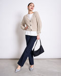 A woman stands against a plain white wall, wearing the jan-n-june CHUNKY CARDIGAN LENA IVORY GOTS over a white top, paired with dark blue jeans, silver pointed-toe heels, and holding a black handbag by her side.