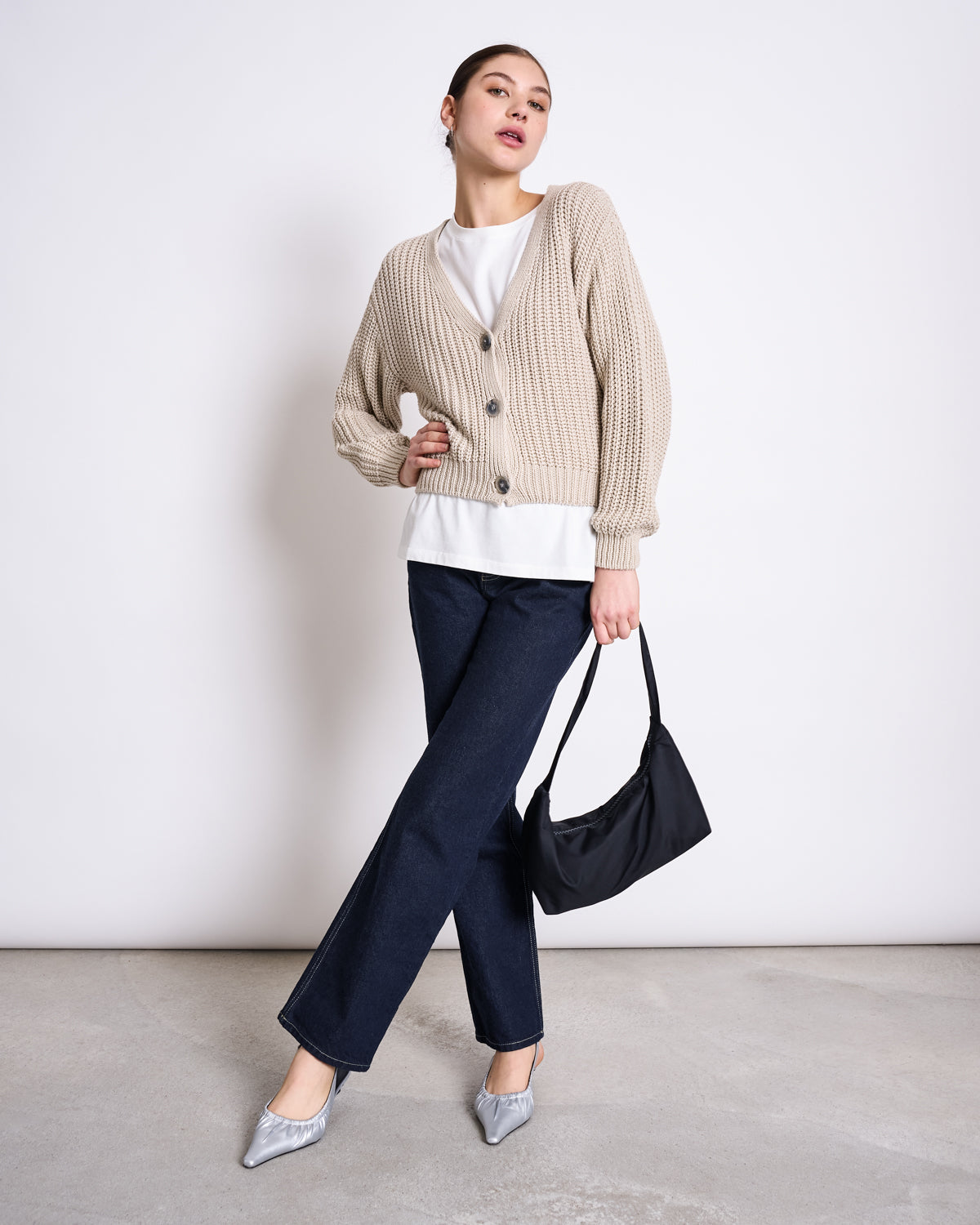 A woman stands against a plain white wall, wearing the jan-n-june CHUNKY CARDIGAN LENA IVORY GOTS over a white top, paired with dark blue jeans, silver pointed-toe heels, and holding a black handbag by her side.