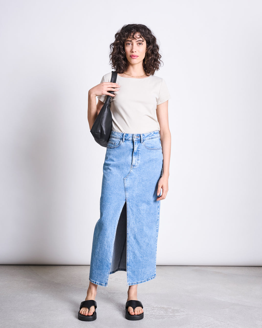 A woman with curly hair wears the jan-n-june BRUSHED BOAT NECK T-SHIRT OTTO IVORY GOTS, a long light blue denim skirt with a front slit, black slide sandals, and carries a black shoulder bag against a plain light background.