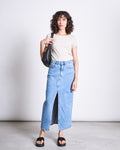 A woman with curly hair wears the jan-n-june BRUSHED BOAT NECK T-SHIRT OTTO IVORY GOTS, a long light blue denim skirt with a front slit, black slide sandals, and carries a black shoulder bag against a plain light background.
