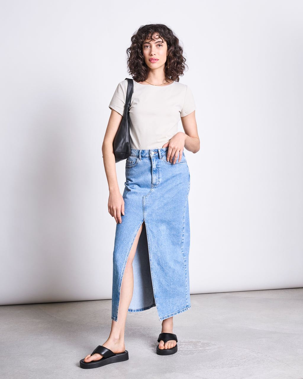 A woman with curly hair and a relaxed pose stands against a plain background, wearing the jan-n-june BRUSHED BOAT NECK T-SHIRT OTTO IVORY GOTS, a long blue denim skirt with a front slit, black sandals, and carrying a black shoulder bag.