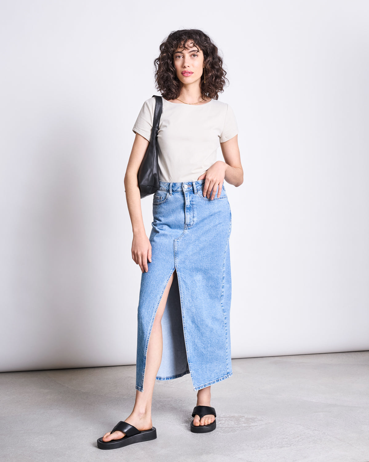 A woman with curly hair and a relaxed pose stands against a plain background, wearing the jan-n-june BRUSHED BOAT NECK T-SHIRT OTTO IVORY GOTS, a long blue denim skirt with a front slit, black sandals, and carrying a black shoulder bag.