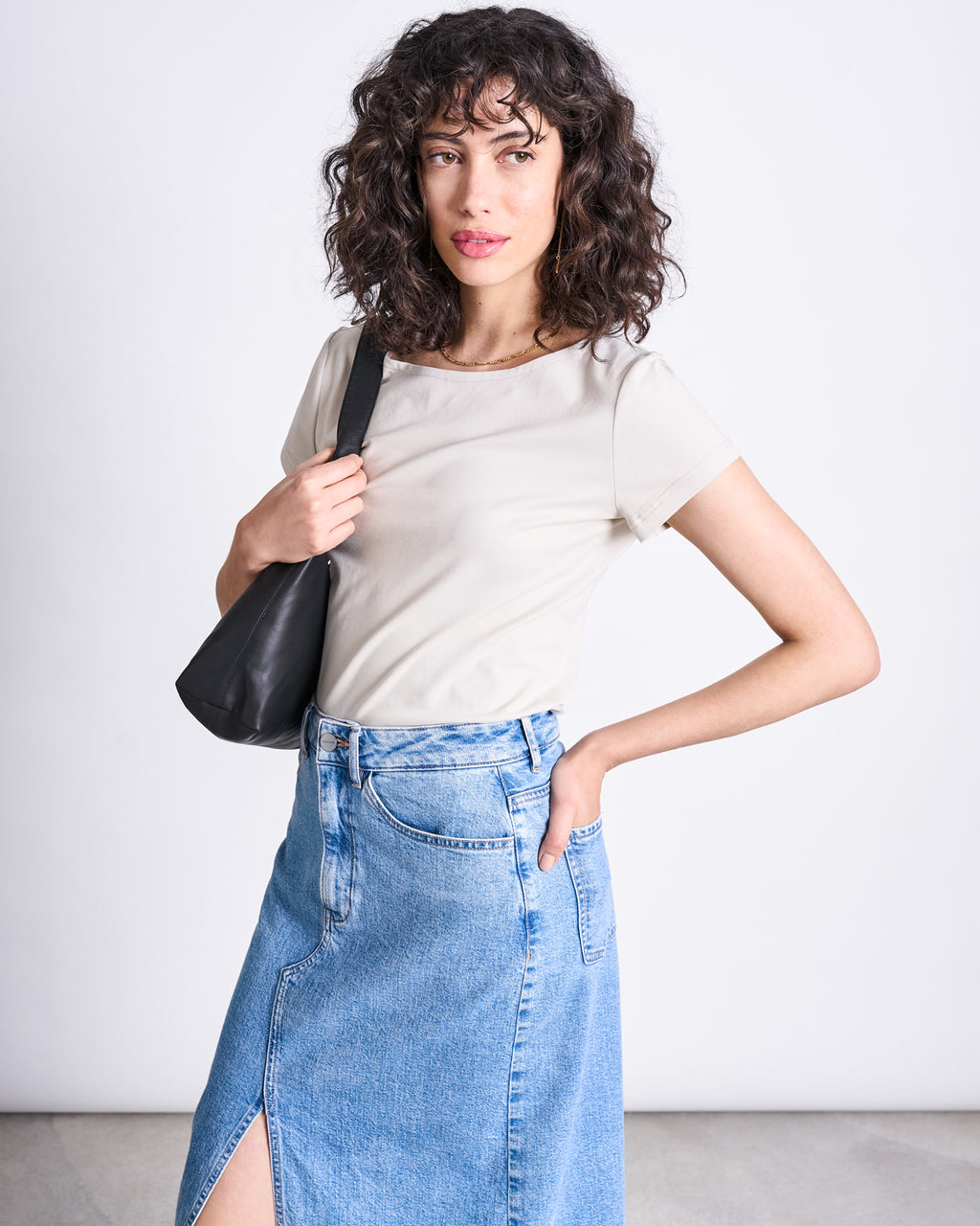 A woman with curly hair wears the jan-n-june BRUSHED BOAT NECK T-SHIRT OTTO IVORY GOTS, paired with a blue denim skirt and a black shoulder bag, posing against a plain light background with one hand in her pocket.