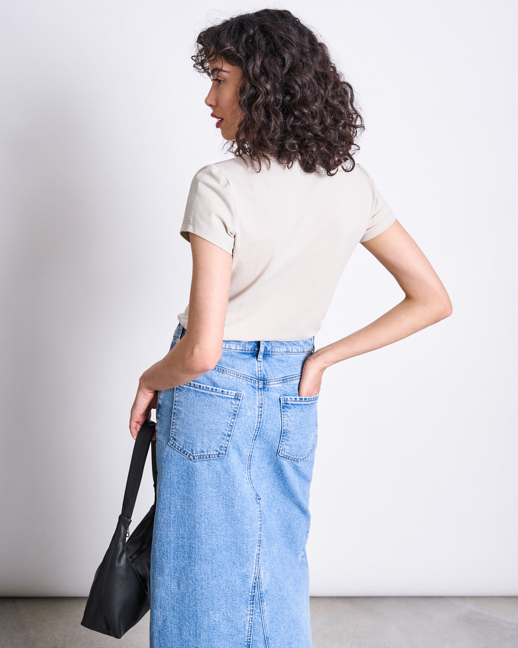 A woman with curly hair faces away, wearing the jan-n-june BRUSHED BOAT NECK T-SHIRT OTTO IVORY GOTS and a high-waisted blue denim skirt. She holds a black bag in her left hand, her right on her waist, against a plain background.