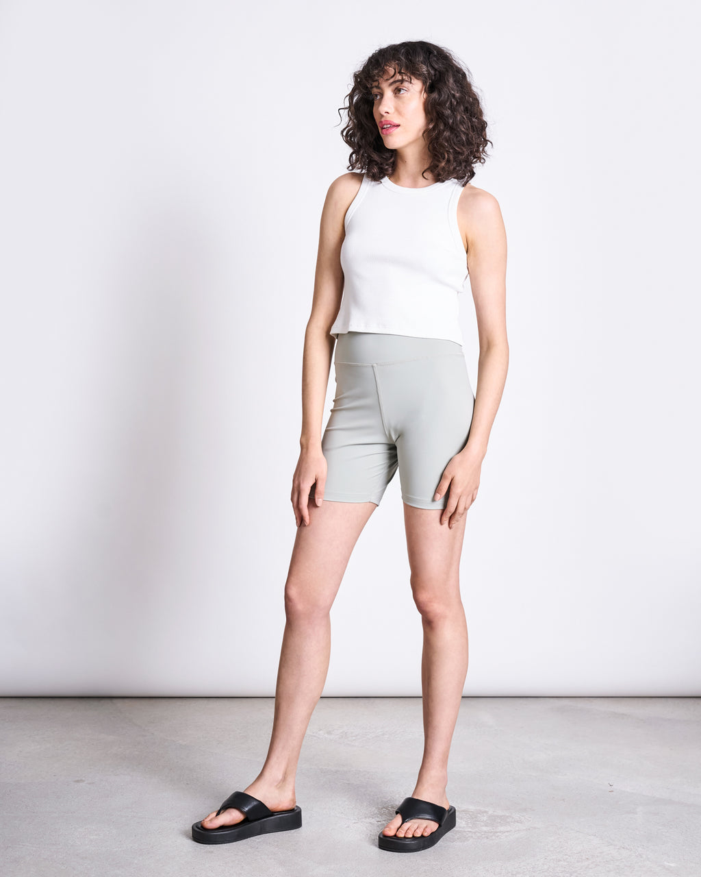 A woman with curly hair wears a white sleeveless top, jan-n-june ACTIVE BIKER SHORTS CRAIL in Glacial Green, and black slide sandals while standing on a concrete floor against a plain white background.