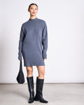 A person stands against a plain background wearing the jan-n-june KNIT DRESS NUNA TURBULENCE GOTS, styled with knee-high black boots and a black bag. Their hair is pulled back, and they have a subtle, neutral expression.