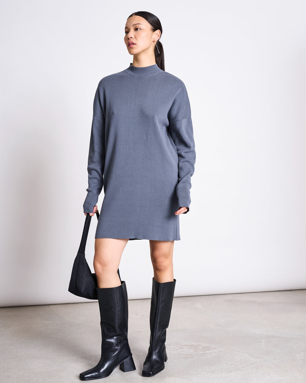 A woman stands against a plain light background wearing the jan-n-june KNIT DRESS NUNA TURBULENCE GOTS in grey with black knee-high boots. She holds a black handbag, and her hair is styled in a sleek ponytail.