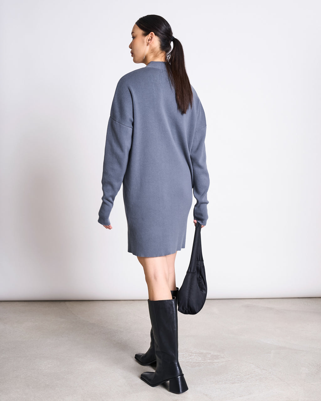 A woman with a low ponytail wears the jan-n-june KNIT DRESS NUNA TURBULENCE GOTS in gray, styled with black knee-high boots and a small black bag, standing against a minimalistic background.