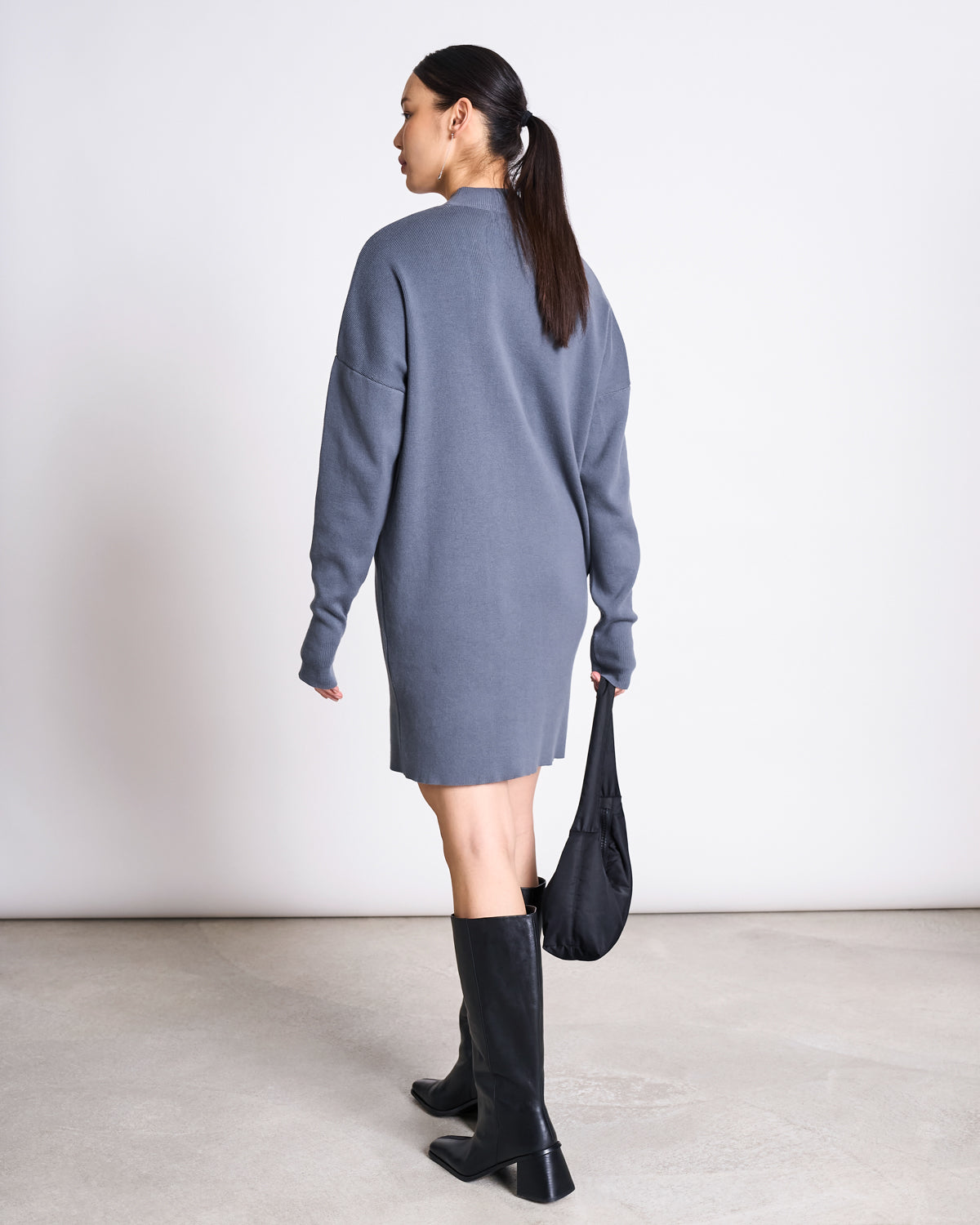 A woman with a low ponytail wears the jan-n-june KNIT DRESS NUNA TURBULENCE GOTS in gray, styled with black knee-high boots and a small black bag, standing against a minimalistic background.